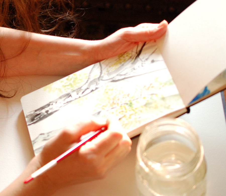 Watercolor sketchbook with a watercolor birch tree. Sunny and inspiring artwork done by hand with an impressionistic style. The sketchbook lays open and we can see the painting from the side. The female painter holds a red paintbrush in her right hand. 